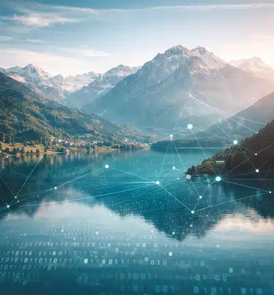 Espace de travail d'un webdesigner freelance installé sur une terrasse en bois. Deux écrans affichent des sites internet "Local Chablais". La vue panoramique donne sur le Lac Léman et les montagnes au lever du soleil. Un carnet de croquis et une tasse à café sont sur le bureau.